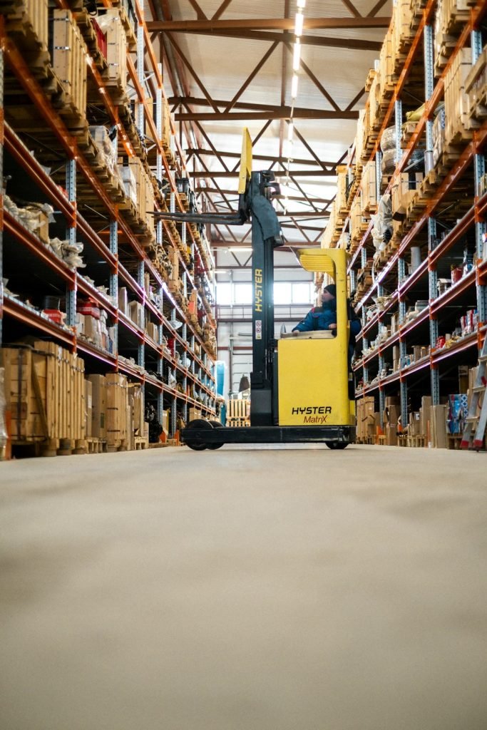 Warehouse Worker Organizing Inventory on a Ladder A warehouse worker in blue overalls and a beanie is standing on a metal ladder, reaching for a package on a high shelf. The warehouse is filled with industrial shelves stocked with boxes and various supplies. The image captures the essence of logistics, inventory management, and manual labor in a structured warehouse environment.